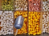 An array of neatly arranged, colorful food items in bins, showcasing various types of goods such as green striped gourd-like vegetables, beige-white round shapes, small reddish cylinders, cubed white items, bright yellow cube-shaped and spherical objects. A metal scoop is resting in one bin, indicating these may be items for sale in a market or store.