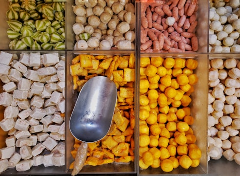 An array of neatly arranged, colorful food items in bins, showcasing various types of goods such as green striped gourd-like vegetables, beige-white round shapes, small reddish cylinders, cubed white items, bright yellow cube-shaped and spherical objects. A metal scoop is resting in one bin, indicating these may be items for sale in a market or store.