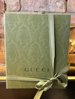 A textured green gift box with an embossed pattern, tied with a matching ribbon that has the word 'GUCCI' printed on it. The box is set against a rustic brick wall, adding an elegant and sophisticated touch.