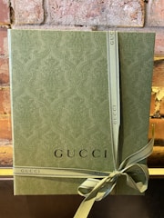 A textured green gift box with an embossed pattern, tied with a matching ribbon that has the word 'GUCCI' printed on it. The box is set against a rustic brick wall, adding an elegant and sophisticated touch.