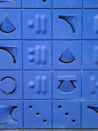 A series of blue blocks featuring various geometric shapes and patterns are displayed. The shapes include triangles, circles, arches, and lines carved into the blocks. The blocks are arranged in a grid pattern, creating a visually pleasing design with textured surfaces.