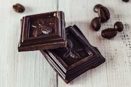 Two pieces of dark chocolate with cacao pod designs rest on a light wooden surface, accompanied by scattered coffee beans.