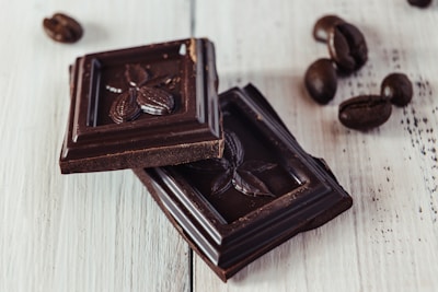Two pieces of dark chocolate with cacao pod designs rest on a light wooden surface, accompanied by scattered coffee beans.