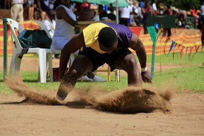 An athlete in a dark shirt and yellow bib is captured mid-action during a long jump event, creating a dramatic spray of sand as they land. The scene is set in an outdoor track and field venue, with spectators and colorful flags in the background adding to the lively atmosphere.