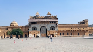 A large open courtyard in front of a historic fort structure with ornate gates and domed towers, featuring a few people walking and taking pictures. The architecture includes intricate carvings and yellowish sandstone walls, with some trees and motorcycles visible on the left side.