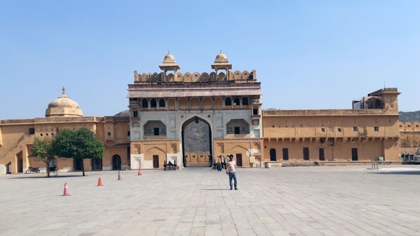 A large open courtyard in front of a historic fort structure with ornate gates and domed towers, featuring a few people walking and taking pictures. The architecture includes intricate carvings and yellowish sandstone walls, with some trees and motorcycles visible on the left side.