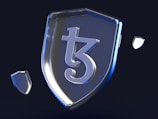 A metallic shield prominently featuring a stylized symbol in the center, with smaller shield-like shapes floating around it against a dark background. The surface of the shield and surrounding elements reflect a blueish tint, giving a modern and sleek appearance.