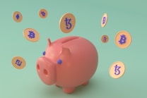 A pink piggy bank surrounded by floating digital currency coins with various cryptocurrency symbols. The background is a soft green color, and the coins feature symbols for Bitcoin, Ethereum, and Binance Coin, among others.