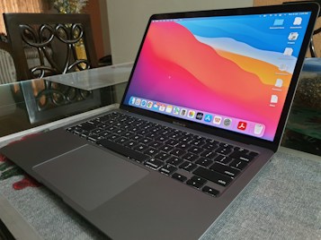 A sleek laptop with a vibrant screen showing a colorful abstract wallpaper, placed on a glass table. The keyboard and trackpad are visible, and the device appears to be a MacBook Air. In the background, part of a decorative chair and a blurred plant arrangement can be seen.