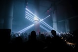 A large crowd gathers in a dark, industrial-looking venue with tall concrete pillars. Bright white spotlights shine from above, creating beams of light that cast over the audience. The atmosphere suggests a lively and energetic event, possibly a concert or a club gathering.