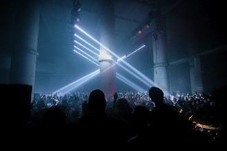 A large crowd gathers in a dark, industrial-looking venue with tall concrete pillars. Bright white spotlights shine from above, creating beams of light that cast over the audience. The atmosphere suggests a lively and energetic event, possibly a concert or a club gathering.