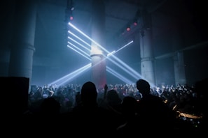 A large crowd gathers in a dark, industrial-looking venue with tall concrete pillars. Bright white spotlights shine from above, creating beams of light that cast over the audience. The atmosphere suggests a lively and energetic event, possibly a concert or a club gathering.