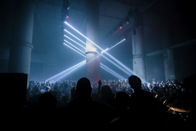 A large crowd gathers in a dark, industrial-looking venue with tall concrete pillars. Bright white spotlights shine from above, creating beams of light that cast over the audience. The atmosphere suggests a lively and energetic event, possibly a concert or a club gathering.