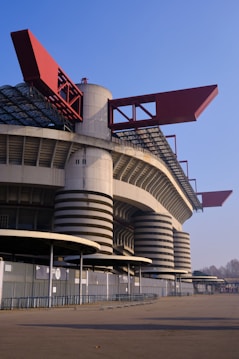 A large, modern stadium with a distinct architectural design featuring cylindrical towers and red rectangular elements. The structure is mostly concrete with black horizontal lines, and the area around it is expansive and empty, suggesting a quiet moment with no events taking place.