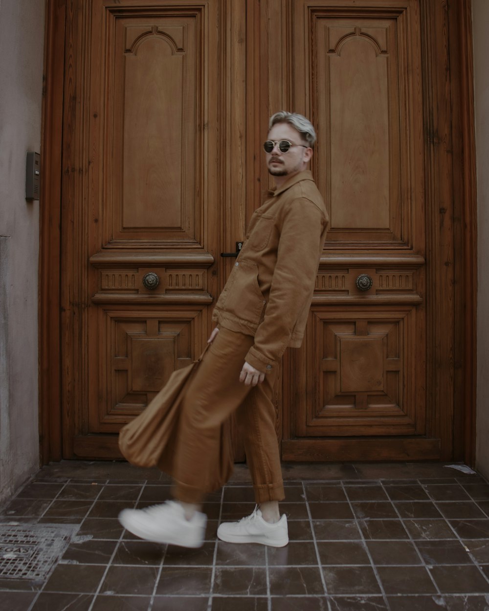 a man in a brown coat and white shoes is standing in front of a door