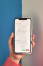 A hand holding a smartphone displaying a cryptocurrency trading application. The screen shows a candlestick chart for BTC/BUSD with a price of 36,437.00, as well as other information like the order book, market history, and margin data. The background has a two-tone color scheme with blue on the left and light gray on the right. The person is wearing a pinkish-red long-sleeve shirt.