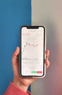 A hand holding a smartphone displaying a cryptocurrency trading application. The screen shows a candlestick chart for BTC/BUSD with a price of 36,437.00, as well as other information like the order book, market history, and margin data. The background has a two-tone color scheme with blue on the left and light gray on the right. The person is wearing a pinkish-red long-sleeve shirt.