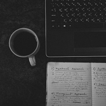 Black and white photo of a person writing notes beside a laptop and a cup of coffee.
