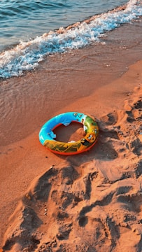 A colorful inflatable swim ring lies on the sandy beach, with gentle waves lapping at the shore. The sand has footprints and small rocks scattered around.