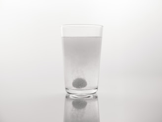 A clear glass filled with water stands on a reflective surface. A spherical tablet is dissolving at the bottom of the glass, releasing bubbles. The background is plain and neutral, highlighting the simplicity of the setting.