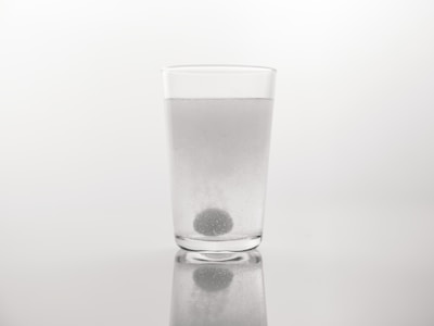 A clear glass filled with water stands on a reflective surface. A spherical tablet is dissolving at the bottom of the glass, releasing bubbles. The background is plain and neutral, highlighting the simplicity of the setting.