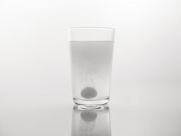 A clear glass filled with water stands on a reflective surface. A spherical tablet is dissolving at the bottom of the glass, releasing bubbles. The background is plain and neutral, highlighting the simplicity of the setting.