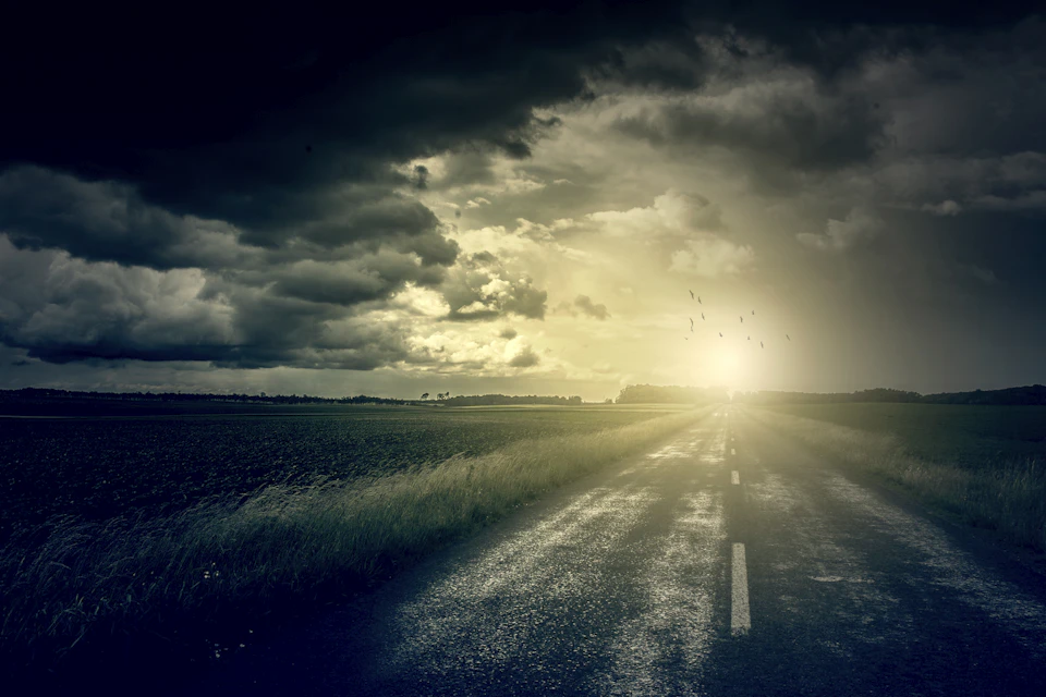 Rural road winding through grassy field under overcast sky with dark clouds and trees