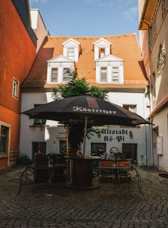 A quaint courtyard surrounded by buildings with distinct red-orange roof tiles and white facades, featuring several stacked chairs beneath a large black patio umbrella with 'Köstritzer' branding. The cobblestone ground adds to the rustic charm, with a lush green plant growing against one of the walls.