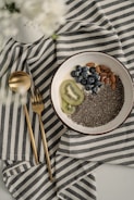 A bowl containing chia seeds, almonds, blueberries, and slices of kiwi rests on a striped fabric cloth. Next to the bowl are a gold-colored spoon and fork. The overall setting is minimalistic and elegant, with a soft focus on white flowers in the top left corner.