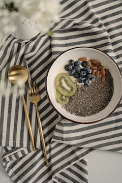 A bowl containing chia seeds, almonds, blueberries, and slices of kiwi rests on a striped fabric cloth. Next to the bowl are a gold-colored spoon and fork. The overall setting is minimalistic and elegant, with a soft focus on white flowers in the top left corner.