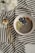 A bowl containing chia seeds, almonds, blueberries, and slices of kiwi rests on a striped fabric cloth. Next to the bowl are a gold-colored spoon and fork. The overall setting is minimalistic and elegant, with a soft focus on white flowers in the top left corner.