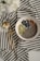 A bowl containing chia seeds, almonds, blueberries, and slices of kiwi rests on a striped fabric cloth. Next to the bowl are a gold-colored spoon and fork. The overall setting is minimalistic and elegant, with a soft focus on white flowers in the top left corner.