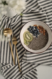 A bowl containing chia seeds, almonds, blueberries, and slices of kiwi rests on a striped fabric cloth. Next to the bowl are a gold-colored spoon and fork. The overall setting is minimalistic and elegant, with a soft focus on white flowers in the top left corner.