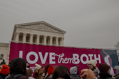 A group of people stand in front of a large pink banner with the phrase 'LOVE them BOTH' displayed in white letters. The setting appears to be an outdoor location with an impressive building in the background, characterized by tall columns and intricate architecture. The crowd is wearing winter clothing, suggesting cold weather.