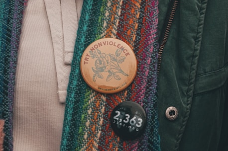 A close-up of two buttons pinned to a multicolored, textured fabric. The upper button is wooden with a design of a rose and the words 'TRY NONVIOLENCE' and 'REHUMANIZEINTL.ORG'. The lower button is black with '2,363 2363.ORG' in white text.