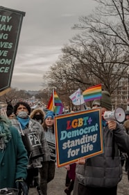 A crowd of people marching in an outdoor protest. Participants hold signs with messages supporting LGBT+ rights. The scene is overcast, and many individuals are dressed warmly. Rainbow flags and banners are visible, expressing solidarity and advocacy.