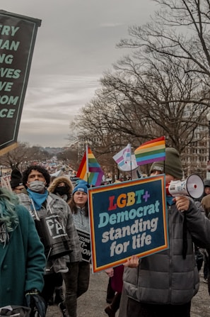 A crowd of people marching in an outdoor protest. Participants hold signs with messages supporting LGBT+ rights. The scene is overcast, and many individuals are dressed warmly. Rainbow flags and banners are visible, expressing solidarity and advocacy.