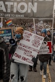 A group of people are participating in a protest or march, holding various signs and banners. One prominent sign reads 'People Over Profit, Babies Over Bottom Lines, Stop the Abortion Industrial Complex, Fund Families.' The crowd includes individuals of different ages, some wearing winter clothing. A rainbow flag is visible in the background.