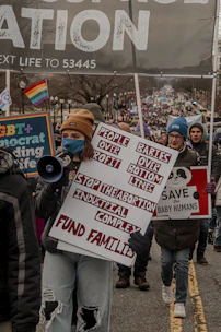 A group of people are participating in a protest or march, holding various signs and banners. One prominent sign reads 'People Over Profit, Babies Over Bottom Lines, Stop the Abortion Industrial Complex, Fund Families.' The crowd includes individuals of different ages, some wearing winter clothing. A rainbow flag is visible in the background.