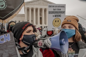 A crowd is gathered in front of a government building, with individuals holding signs related to a protest. One person is speaking into a megaphone, while others hold signs with anti-abortion messages. The participants are wearing winter clothing, such as hats and scarves. The architecture of the building is classical, with large columns.
