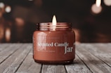 Close-up of a softly glowing scented jar candle with a minimalist label on a wooden table.