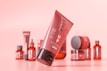 A collection of cosmetic products with a modern packaging design, predominantly in tones of red and pink. The main focus is on a tilted tube with the word 'Mockup' visible, and various other bottles and jars are blurred in the background.