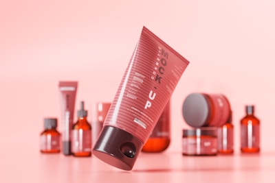 A collection of cosmetic products with a modern packaging design, predominantly in tones of red and pink. The main focus is on a tilted tube with the word 'Mockup' visible, and various other bottles and jars are blurred in the background.