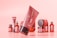 A collection of cosmetic products with a modern packaging design, predominantly in tones of red and pink. The main focus is on a tilted tube with the word 'Mockup' visible, and various other bottles and jars are blurred in the background.