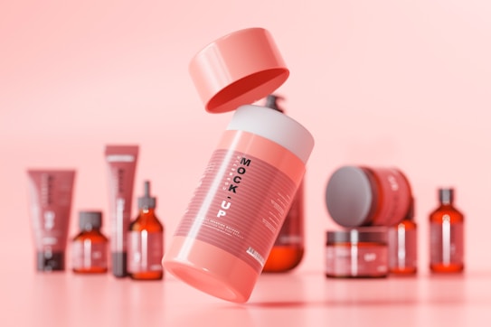 A group of cosmetic containers, including bottles, tubes, and jars, are arranged with a floating bottle in the center. The image has a pink background and the containers vary in shades of red and pink, with labels that read 'MOCK UP'. The composition is neatly organized for a product display.