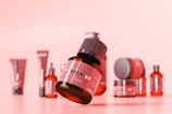 A collection of various cosmetic or skincare product containers arranged on a soft pink background. The central focus is a brown bottle with a black cap, labeled 'MOCK-UP', appearing to be floating or elevated above the surface. Around it, there are other bottles, jars, and tubes in different sizes and shapes, all maintaining a consistent design theme with red and black color schemes.
