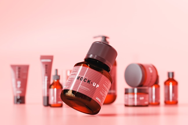 A collection of various cosmetic or skincare product containers arranged on a soft pink background. The central focus is a brown bottle with a black cap, labeled 'MOCK-UP', appearing to be floating or elevated above the surface. Around it, there are other bottles, jars, and tubes in different sizes and shapes, all maintaining a consistent design theme with red and black color schemes.