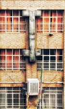 A brick building facade with four large windows, each featuring red curtains. A large, gray architectural structure resembling a cross is centered between the windows. Below the windows, an air conditioning unit is mounted on the wall.