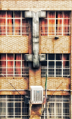A brick building facade with four large windows, each featuring red curtains. A large, gray architectural structure resembling a cross is centered between the windows. Below the windows, an air conditioning unit is mounted on the wall.