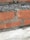 A close-up view of a brick wall segment featuring red bricks and concrete mortar. A noticeable crack runs vertically through one of the bricks, revealing rough and uneven edges. The mortar appears somewhat loose and crumbly in areas.